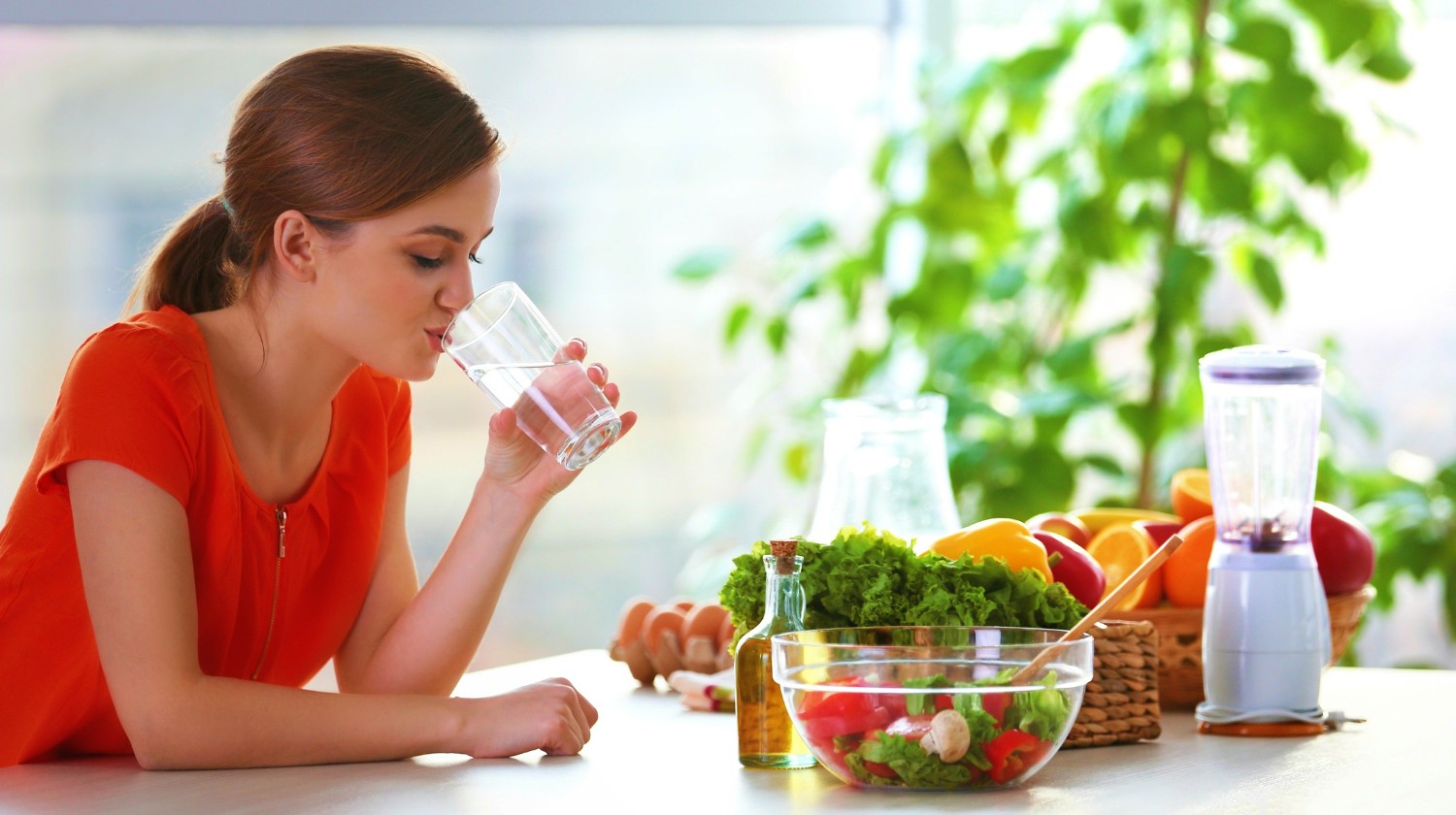 Young woman drinking water near table with fruits and vegetables in the kitchen | High Alkaline Foods To Add To Your Diet